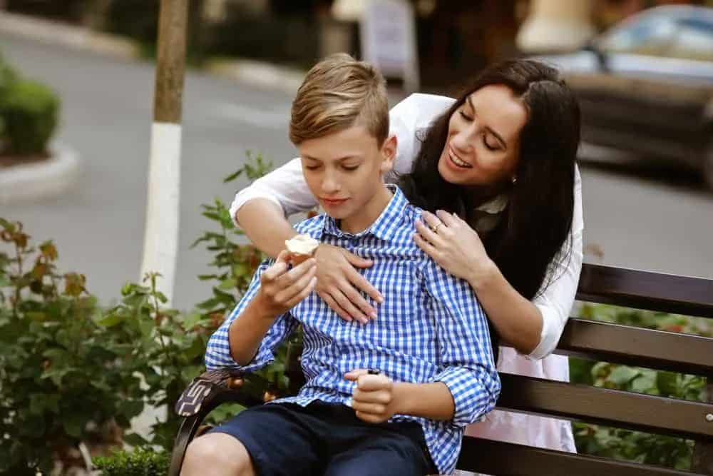 Eine Mutter umarmt ihren Sohn hinter ihrem Rücken, der auf einer Bank sitzt und Eis isst