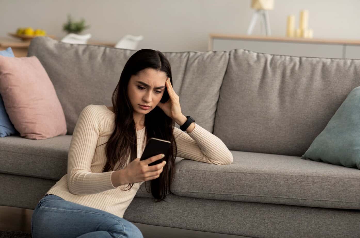 Frau schaut auf ihr Smartphone und sitzt auf dem Boden neben dem Sofa
