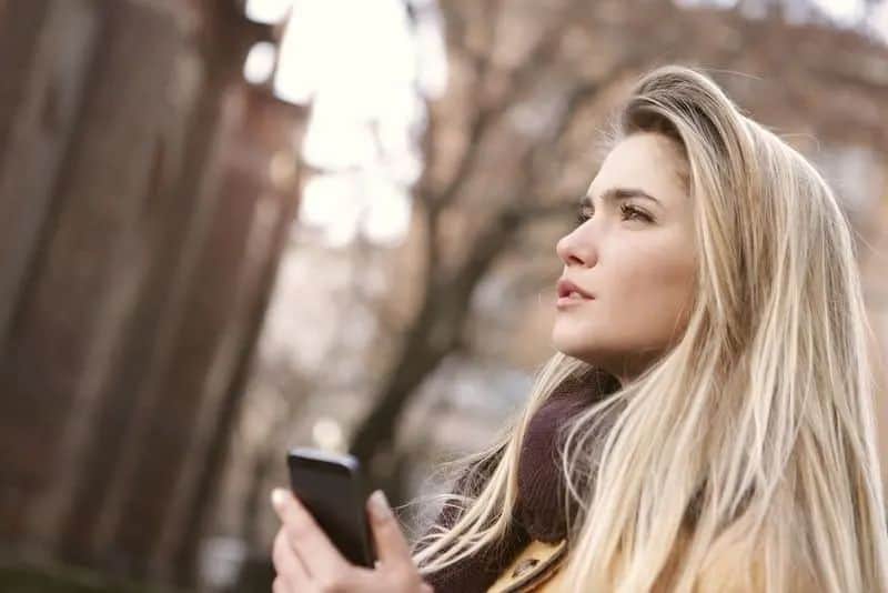Eine nachdenkliche Blondine steht mit einem Telefon in der Hand im Wald