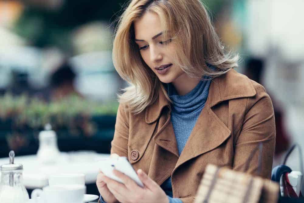 Eine schöne Blondine in einem braunen Mantel sitzt auf der Terrasse eines Cafés und schreibt eine SMS