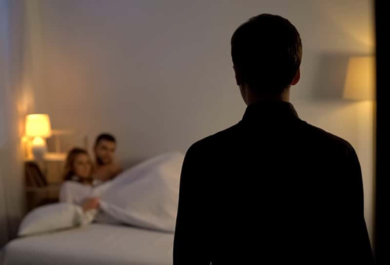 Ein Mann, der seine Freundin im Bett mit einem anderen Mann ansieht, während er vor ihnen steht