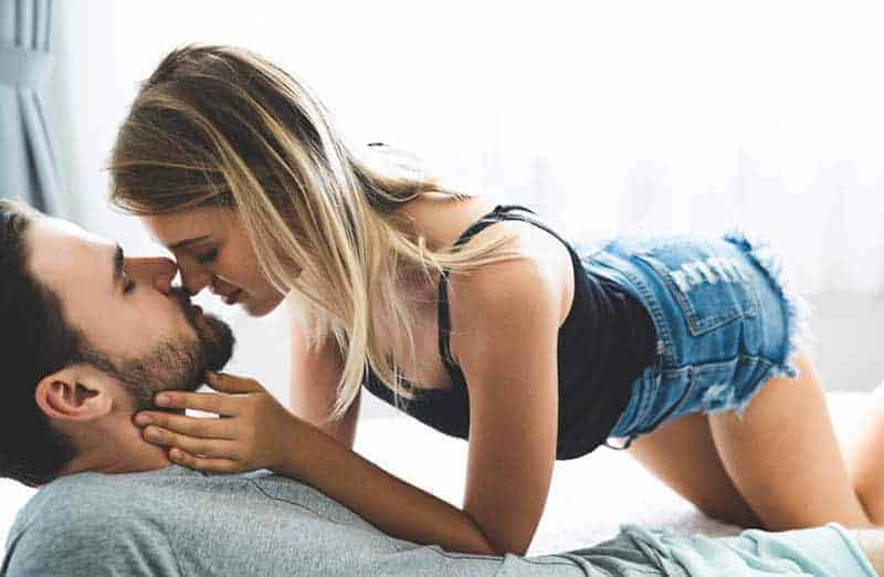 Auf dem Bett küsst eine Frau in Shorts und einem schwarzen T-Shirt einen Mann mit Bart