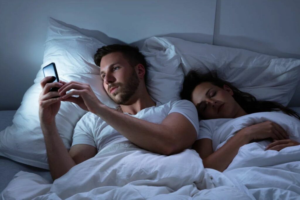 Ein Mann im Bett tippt auf seinem Telefon, waehrend seine Frau schlaeft