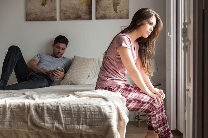 traurige-junge-Frau-sitzt-auf-dem-Bett-während-Mann-SMS-am-Telefon
