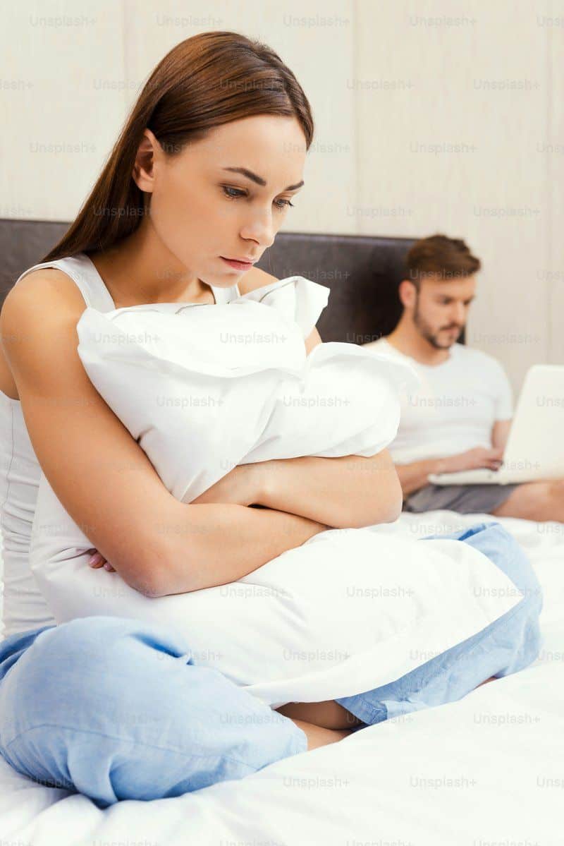 Depressed young woman embracing pillow and looking down while sitting on bed with young man working on laptop in the background photo – Stress Image on Unsplash Depressed young woman embracing pillow and looking down while sitting on bed with young man working on laptop in the background photo – Stress Image on Unsplash