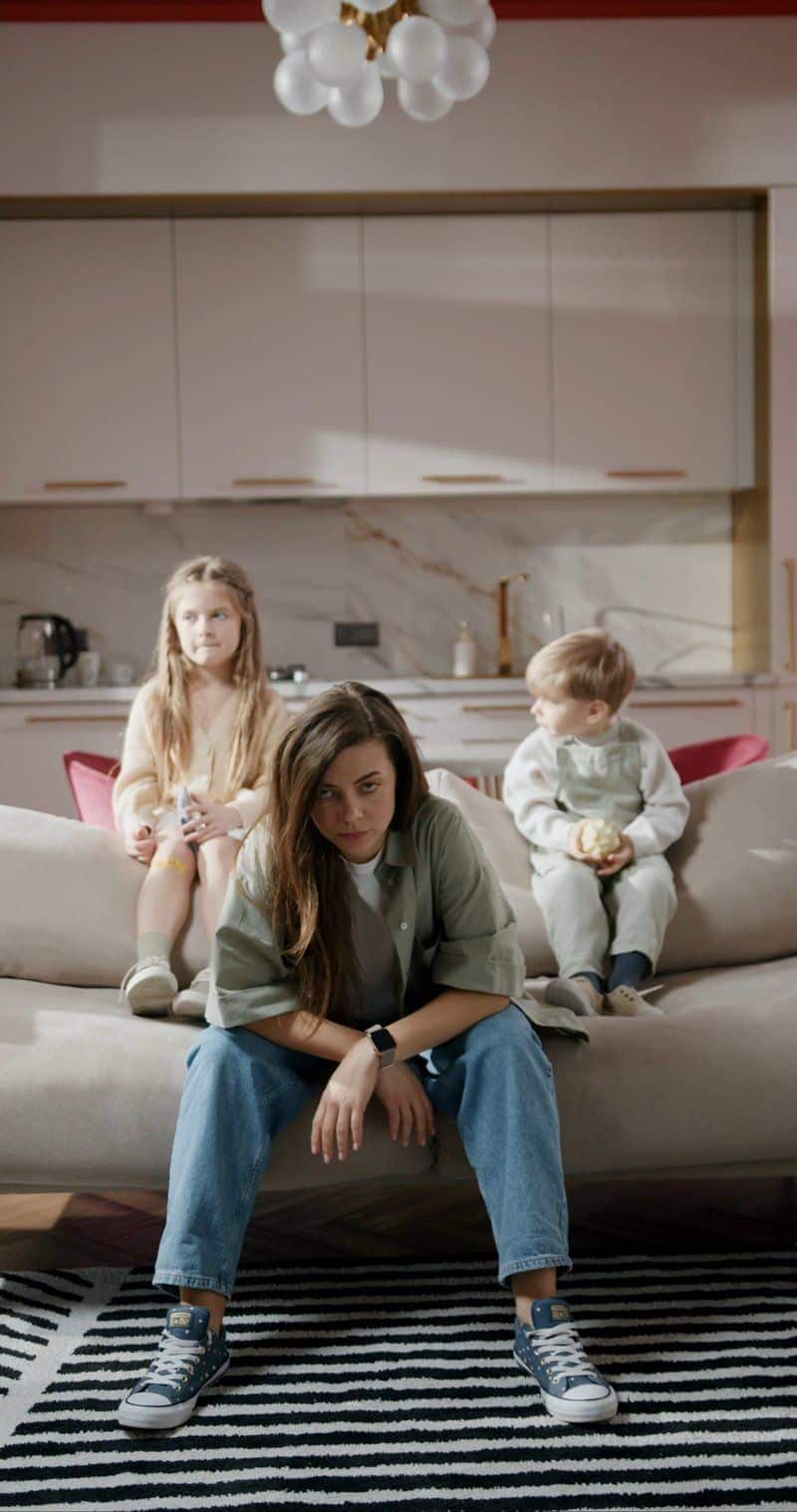 A Mother Feeling Tired While Sitting On Sofa With Her Children A Mother Feeling Tired While Sitting On Sofa With Her Children